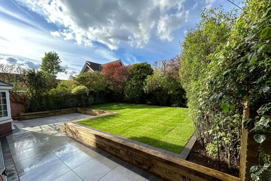 Neatly landscaped backyard with fresh green lawn, wooden retaining walls, stone tiled patio, and surrounding trees under a partly cloudy sky.