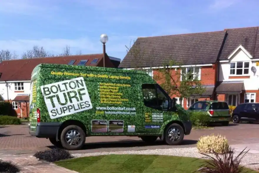 A Bolton Turf Supplies van, adorned with lush turf graphics and company contact details, is parked on a landscaped residential street with houses and gardens visible in the background. The scene includes well-maintained lawns, vibrant plants, and a paved driveway.