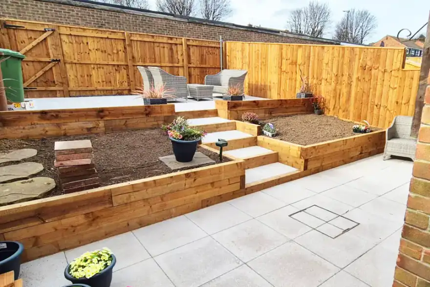 Tiered garden featuring raised timber planting beds, wide paved steps, and a spacious light-grey porcelain patio. A raised seating area with outdoor furniture is enclosed by timber fencing, creating a structured and modern outdoor space.