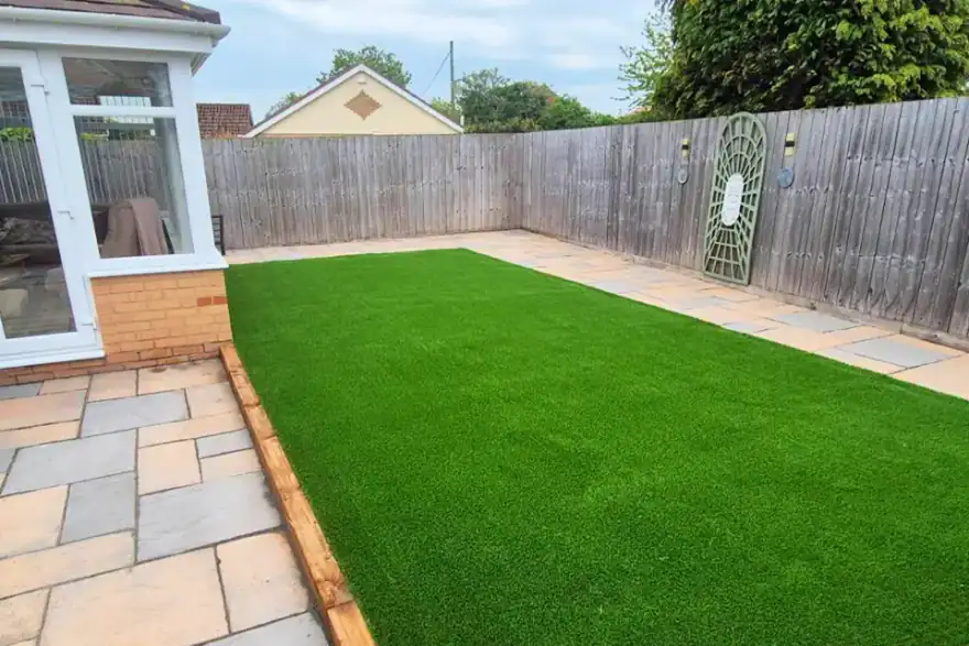 Low-maintenance garden featuring a large artificial grass lawn bordered by natural stone paving. The garden is enclosed by timber fencing and adjoins a conservatory, creating a tidy and practical outdoor living space.