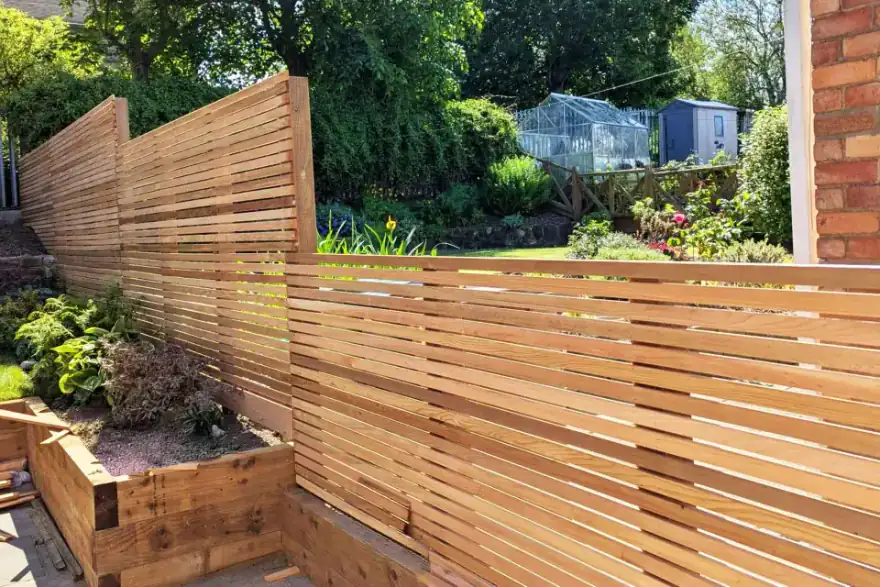 Contemporary timber slatted fencing and raised wooden planters bordering a landscaped garden. Established planting, a greenhouse, and mature trees provide privacy and structure to the outdoor space.