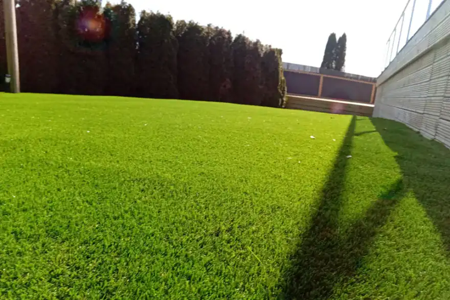 Bright green artificial grass lawn with shadows from a fence on the right and trees in the background, illuminated by sunlight.