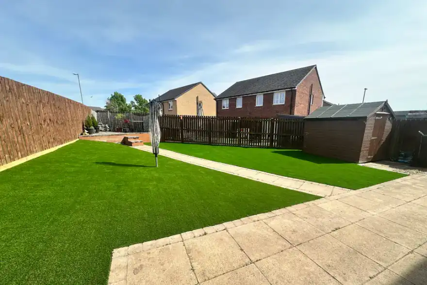 A backyard with a paved patio area, a green artificial grass lawn, a brown shed, and a wooden fence, under a clear blue sky.